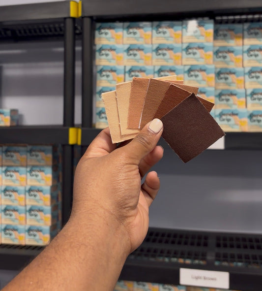 A hand holds a fanned-out set of Gender Grip tape color swatches in front of shelves stocked with matching packaged Gender Grip samples.