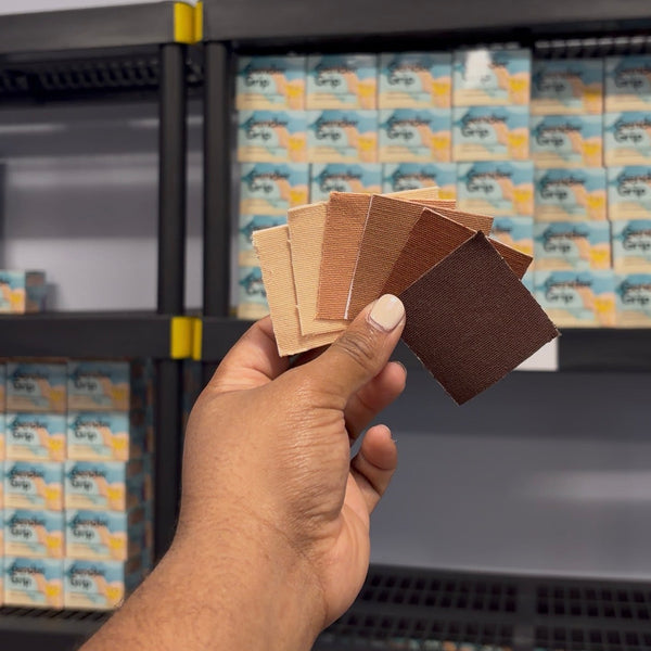 A hand holds a fanned-out set of Gender Grip tape color swatches in front of shelves stocked with matching packaged Gender Grip samples.
